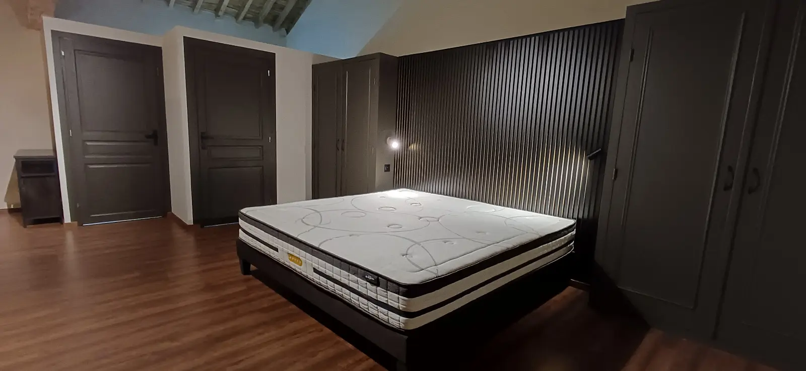 Chambre rénovée : tête de lit en tasseaux de bois sombre, portes et placards peints en anthracite, sol souple imitation parquet.