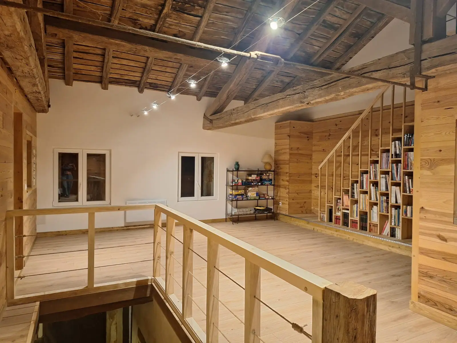 Mezzanine en bois clair sous une charpente ancienne conservée : garde-corps à câbles, escalier bibliothèque, éclairage sur câble tendu.