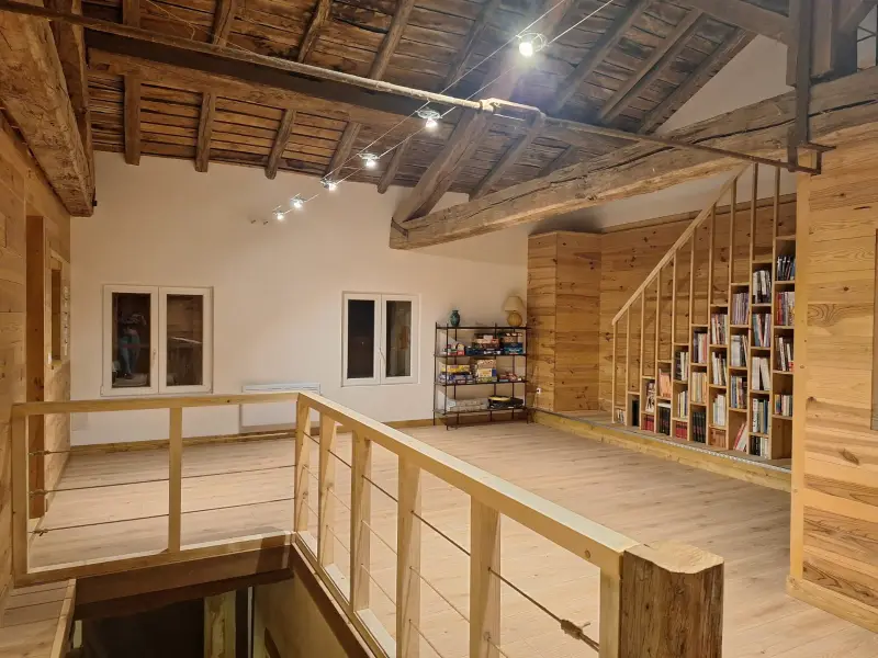 Mezzanine en bois clair sous une charpente ancienne conservée : garde-corps à câbles, escalier bibliothèque, éclairage sur câble tendu.