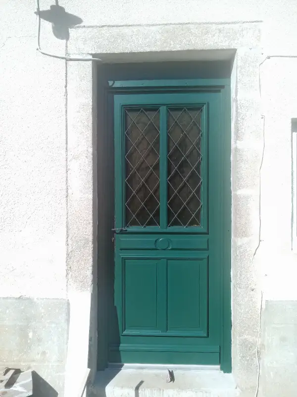 Porte d’entrée en bois peinte en vert, vitrage à petits carreaux, posée dans un encadrement en pierre d’une maison de bourg.