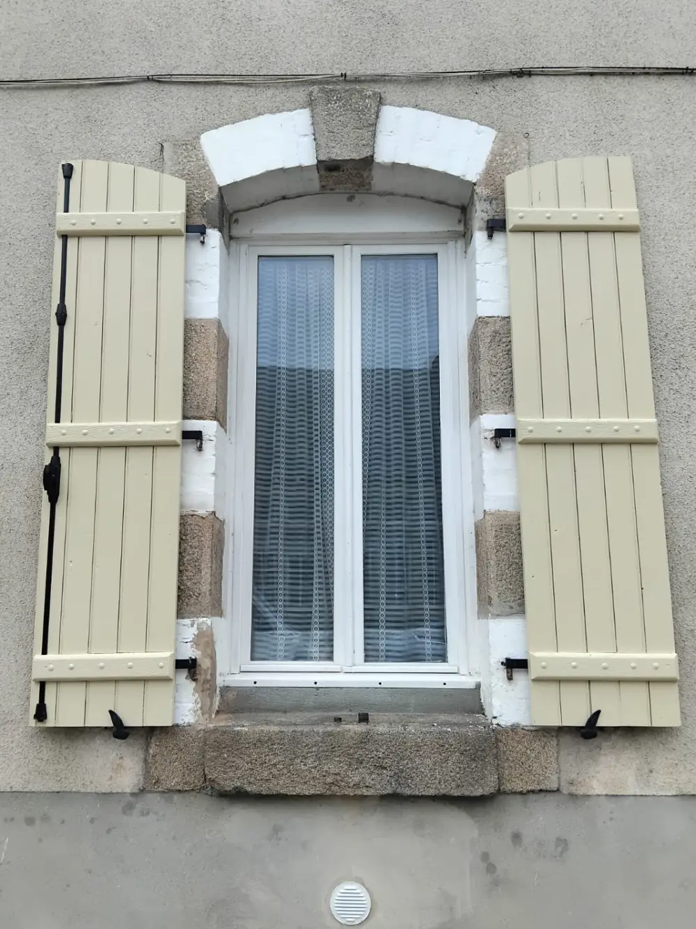 Volets repeints en crème clair, ouverts de part et d’autre de la fenêtre, pentures et arrêts repeints en noir.